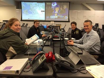 image of members of DEM sitting around a table and smiling at the camera. In the background are large screens displaying maps and many whiteboards