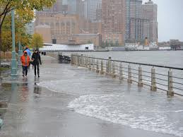 image showing hudson river storm surge flood waters covering most of the surface of a riverfront walkway, with two pedestrians dressed in rain gear walking towards the camera.