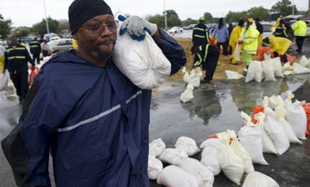 Man standing in the rain wearing a raincoat, with a sandbag on his shoulder. Around him are more sandbags.